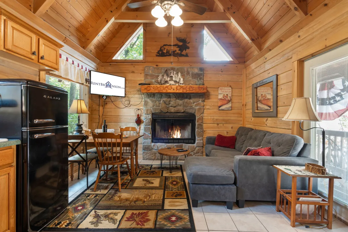 A warm and inviting living space showcasing a classic log fireplace, comfortable sofa, a dining table ideal for meals or games, and a glimpse of the kitchen area. The wood interior and homey décor accentuate both comfort and functionality, with visible amenities that set the stage for movie nights or family gatherings.