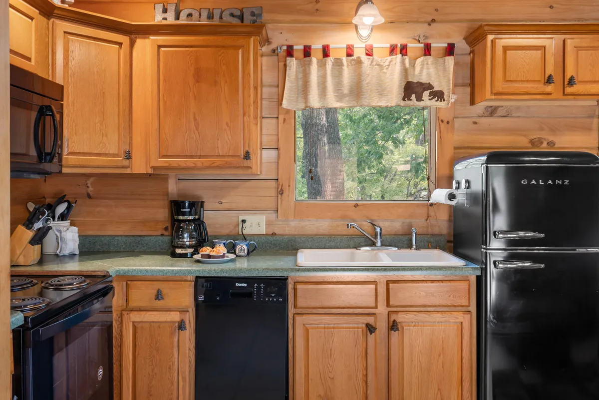 A view of the kitchen area featuring a stove, refrigerator, and sink, with morning light pouring through the window. The layout is functional and inviting, emphasizing the essentials for cooking and socializing around food.