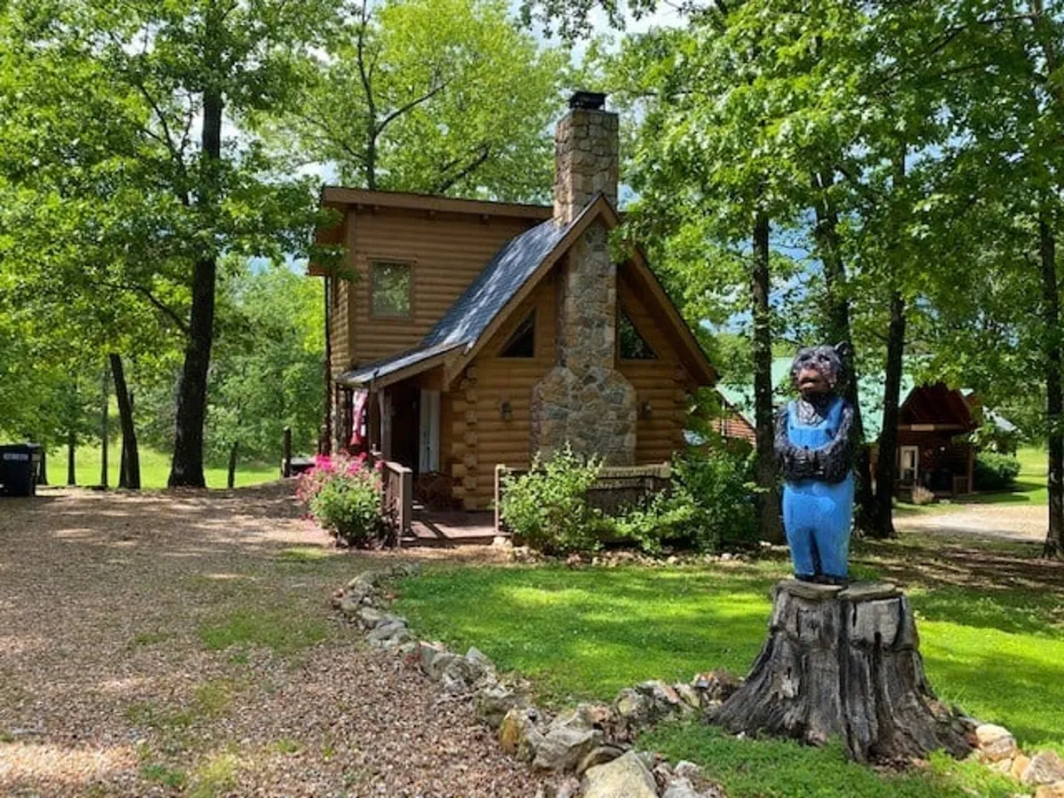 A charming log cabin with a stone chimney, wooden bear statue, and flowering plants on the porch, set amidst tall trees. The inviting porch hints at peaceful mornings, evening barbecues, and easy access to nature trails.