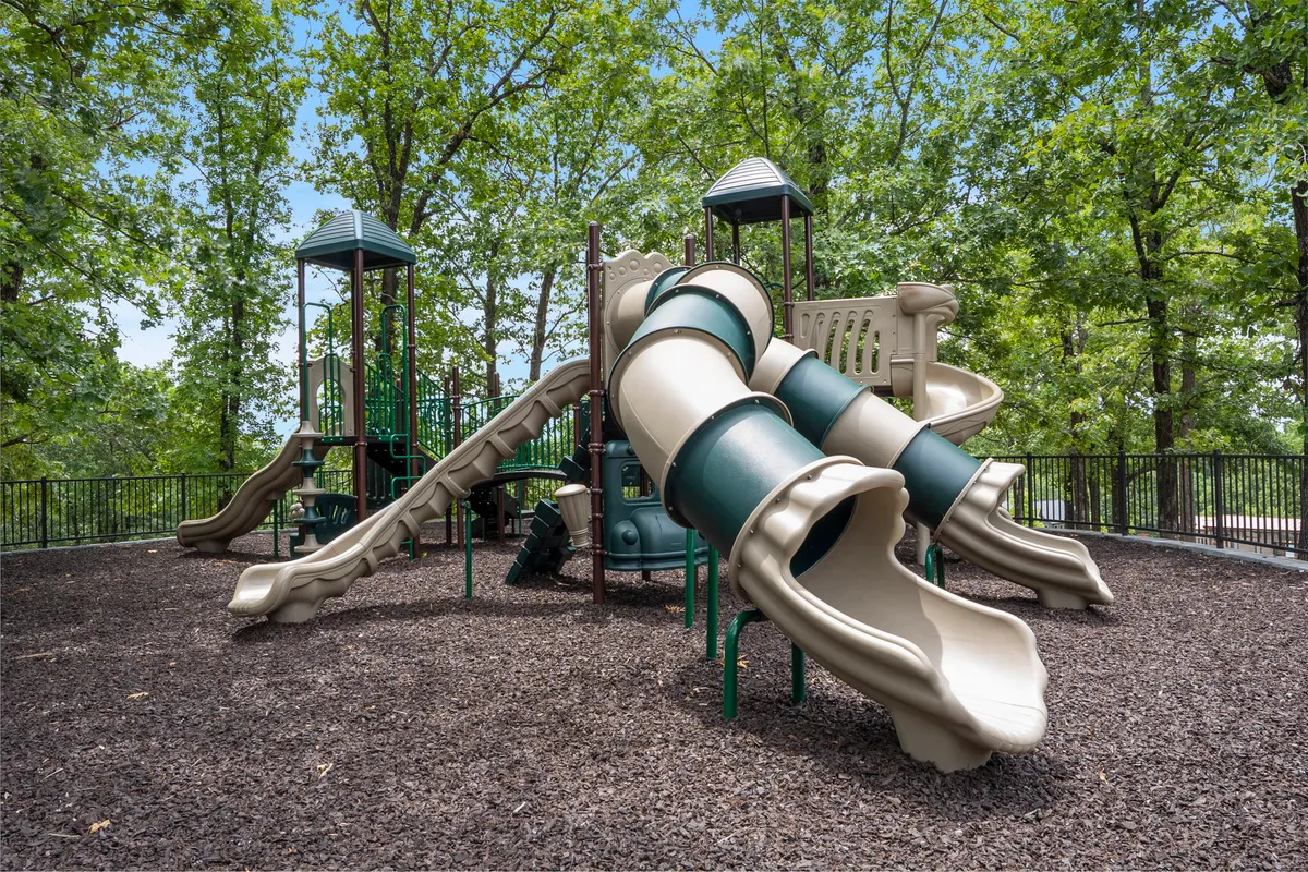 This image features a playground with slides and climbing structures surrounded by trees. Children can be seen actively playing, and there is ample space for family gatherings and picnics, all set within a lush, natural environment.