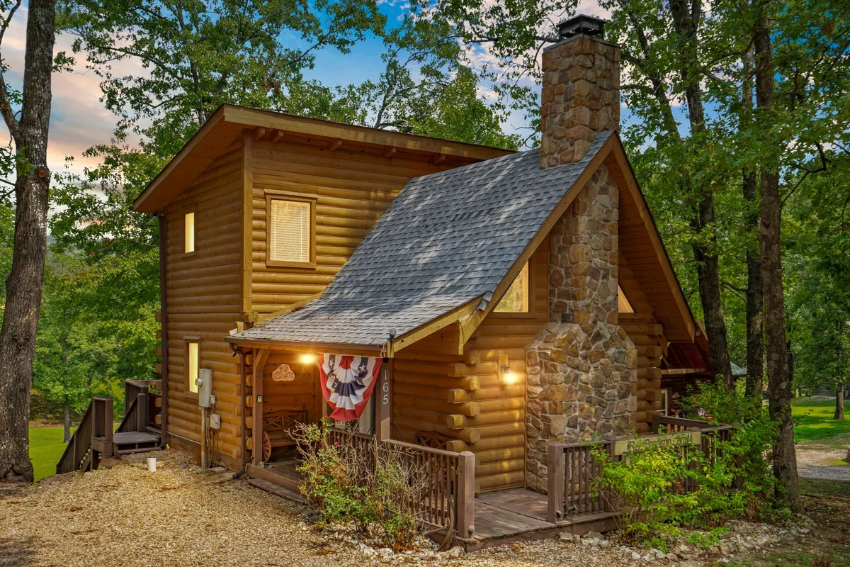 The image showcases a charming, all-wood log cabin with a prominent stone chimney, surrounded by a beautifully landscaped garden. A spacious wooden deck wraps around the cabin, inviting guests to relax and soak in views of the natural surroundings. The lush greenery sets a tranquil mood, perfect for quiet moments or gathering with loved ones.