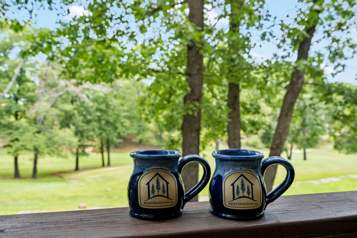 Two blue mugs rest on a wooden railing, framed by lush green trees and a distant, serene landscape. The scene evokes peaceful mornings spent sipping coffee together in nature.