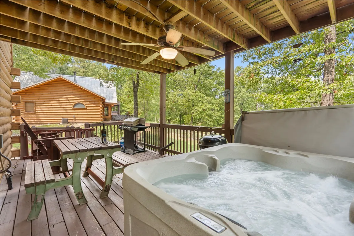 This image highlights a spacious wooden deck surrounded by lush trees, featuring a private hot tub, a gas grill, and a dining area perfect for outdoor meals. The tranquil setting is ideal for relaxing in the hot tub, barbecuing, or enjoying the sunset over the Ozarks.