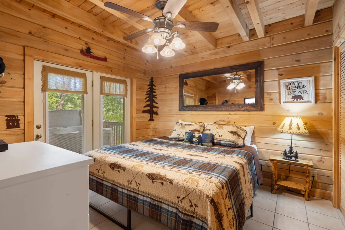 A cozy bedroom featuring a wooden bed frame with a plaid bedspread, a decorative lamp, wooden ceiling, and a mirror. The room is filled with warm, natural light and offers a tranquil atmosphere for relaxation or reading.