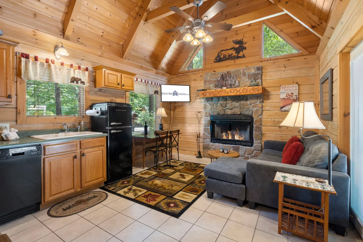 A spacious, warmly lit interior featuring a stone fireplace, plush sofa, dining table, and a view into the well-equipped kitchen. The open concept design is evident, with ample space for both relaxation and socializing, and the overall ambiance is cozy and inviting—perfect for evenings spent indoors.