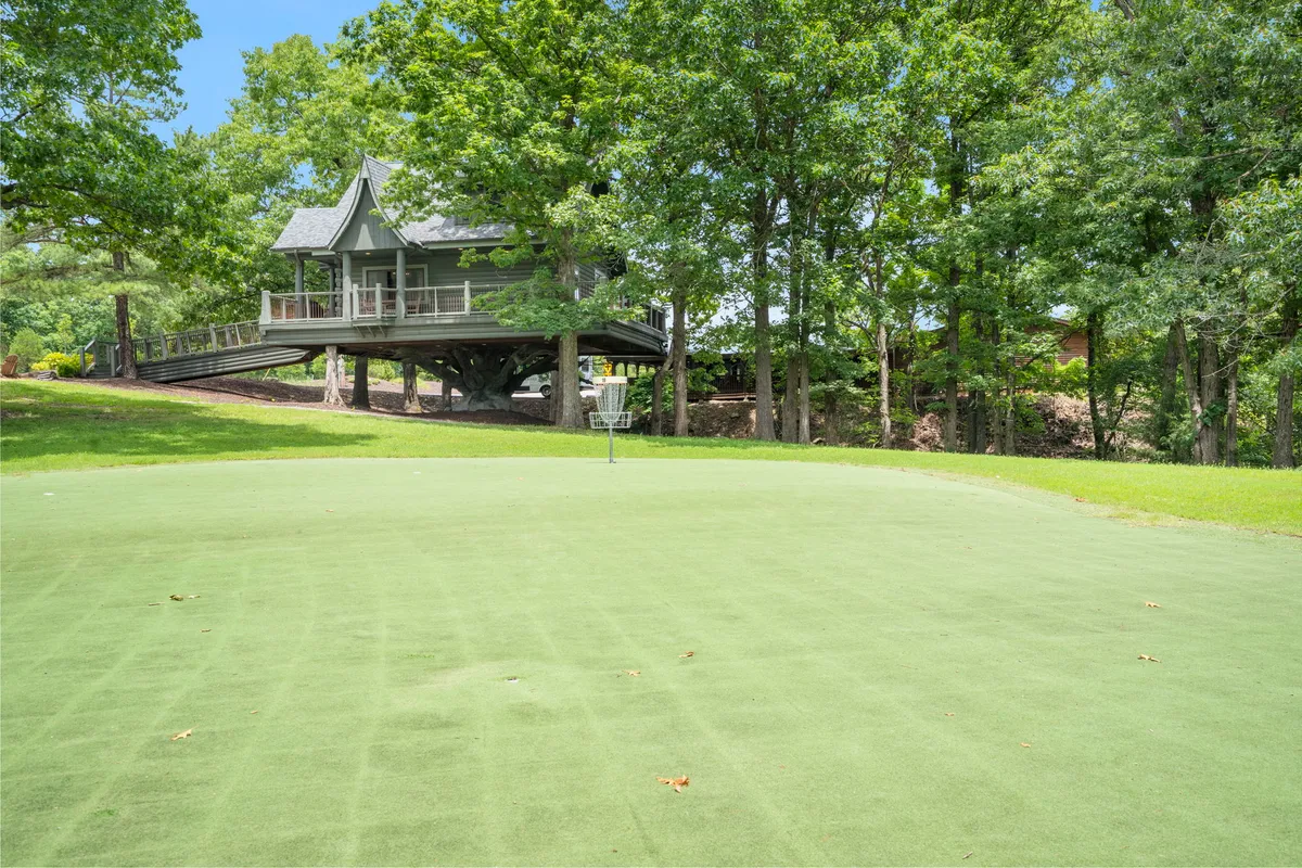 A striking exterior shot showing an elevated house with a manicured putting green out front and tall trees framing the scene. The setting suggests a space for both relaxation and golf practice, with ample greenery and a peaceful, recreational vibe.